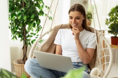 Evden çalışan serbest çalışan, asılmış bir sandalyede oturan, etrafı bitkilerle çevrili bir dizüstü bilgisayarla evinin keyfini çıkaran biri.