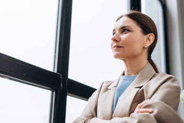 İş kadını, kollarını kavuşturmuş, düşünceli bir şekilde ofisin penceresinden dışarı bakıyor, iş dünyasındaki gelecek fırsatları ve zorlukları düşünüyor.