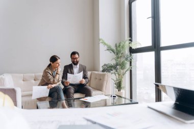 İki gülümseyen iş ortağı modern ofiste koltukta oturup belgeleri analiz etmeyi tartışıyorlar.