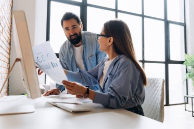 Modern bir ofiste iş birliği yapan meslektaşlar, büyüme stratejilerini tartışırken masaüstü bilgisayarlarında finansal grafikleri analiz ediyorlar.