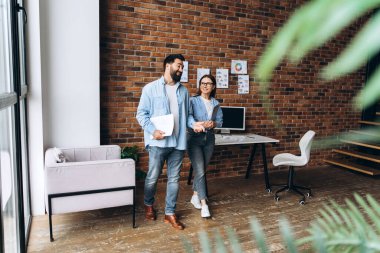 İki meslektaş modern bir ofiste yürürken, etrafı tuğladan duvarlarla çevrili, parlak, havadar bir dekorla çevrili bir şekilde iş konuşuyorlar.