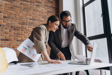 İki mimar, ofislerindeki bir proje üzerinde işbirliği yaparak planlarını ve finansal tablolarını inceliyor.