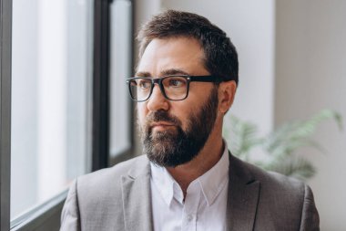 Ofiste takım elbiseli ve gözlüklü bir işadamı pencereden dışarı bakıyor, gelecek fırsatları düşünüyor.