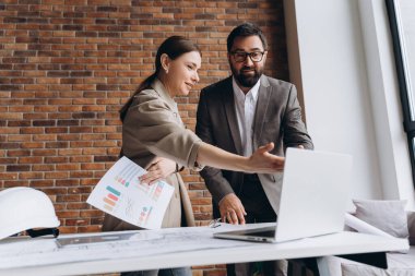 İki mimar bir proje üzerinde işbirliği yapıyor, bir dizüstü bilgisayar kullanırken planları tartışıyor ve ofis alanlarındaki yazılı belgeleri gözden geçiriyorlar.