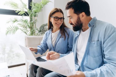 İki iş ortağı bir kanepede oturmuş dizüstü bilgisayardaki bir projeyi ve bazı belgeleri gözden geçiriyorlar.