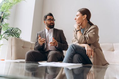 Finansal danışman, kanepede oturan düşünceli müşteriye yatırım stratejisini anlatırken el kol hareketi yapıyor.