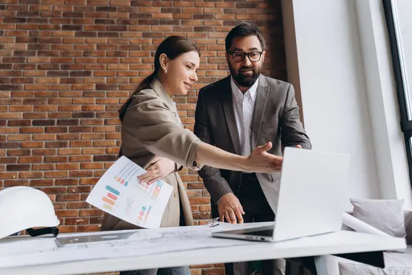 İki mimar bir proje üzerinde işbirliği yapıyor, bir dizüstü bilgisayar kullanırken planları tartışıyor ve ofis alanlarındaki yazılı belgeleri gözden geçiriyorlar.