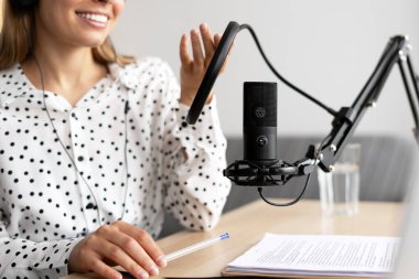 Gülümseyen kadın radyo sunucusu kulaklık takıyor, mikrofona konuşuyor ve modern ses ekipmanları ve dizüstü bilgisayarlarla dolu rahat ev stüdyosunda podcast yapıyor. Çevrimiçi teknoloji kavramı