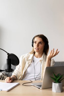 Genç kadın ses içeriğini profesyonel mikrofon ve dizüstü bilgisayar kullanarak kaydediyor, kulaklık takıyor ve konuşurken el kol hareketi yapıyor, evde online içerik yaratıcısı olarak çalışıyor. Canlı, iletişim konsepti
