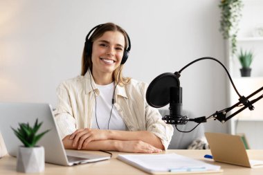 Gülümseyen kadın podcaster kulaklık takarak stüdyoda podcast kaydediyor, kameraya bakarken profesyonel mikrofon ve dizüstü bilgisayar kullanıyor. Canlı, çevrimiçi teknoloji kavramı