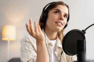 Güzel bir kadın kendi stüdyosunda kulaklık ve mikrofon kullanarak podcast yaparken konuşuyor ve el kol hareketi yapıyor. Canlı, canlı konsept