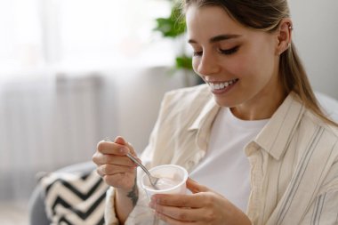Evdeki rahat koltukta oturan genç bir kadın kremalı yoğurdun tadını çıkarırken bir yandan da vücudunu sağlıklı beslenme alışkanlıklarıyla besliyor. Beslenme kavramı