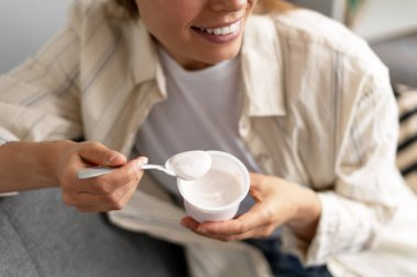 Plastik bardaktan yoğurt yiyen gülümseyen bir kadın, sağlıklı bir atıştırmalık veya kahvaltıdan keyif alan, sağlıklı bir yaşam tarzını teşvik eden. Dengeli beslenme kavramı