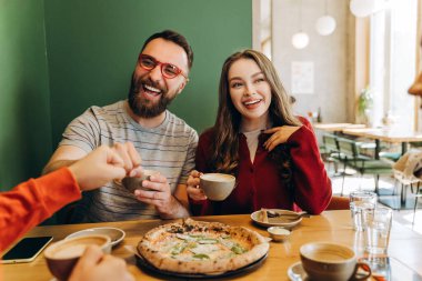 Neşeli arkadaşlar bir restoran masasında yemek yiyorlar, lezzetli pizza ve kahveyi paylaşıyorlar, birlikte gülüyorlar ve neşeli bir arkadaşlık ve bağlılık gösterisi için yumruk tokuşturuyorlar. İletişim konsepti