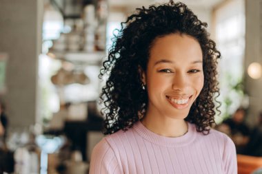 Güzel kıvırcık saçlı, modern bir kahve dükkanında gülümseyen ve poz veren mutlu genç bayan girişimci, iş günü boyunca rahatlatıcı bir anın tadını çıkarıyor.