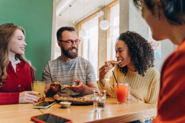 Neşeli çoklu etnik arkadaşlar birlikte pizza yemekten ve meyve suyu içmekten, bir restoranda masada oturmaktan, iyi vakit geçirmekten ve konuşmaktan zevk alıyorlar. Yiyecek konsepti