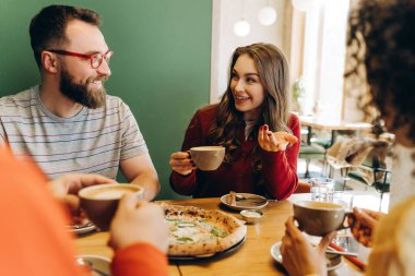 Neşeli bir grup arkadaş, sıcak bir kafede lezzetli pizzanın ve taze demlenmiş kahvenin tadını çıkarıyor, canlı sohbetler yapıyor, gülüyor ve birlikte neşeli anlar paylaşıyorlar. Rahatlatıcı kavram