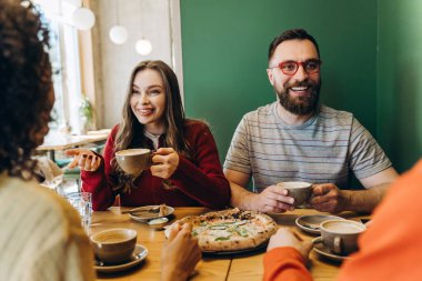 Çeşitli çok uluslu arkadaşlar bir kafede birlikte pizza yiyip kahve içiyorlar, canlı sohbetler yapıyorlar ve sıcak, sosyal bir ortam yaratıyorlar. Arkadaşlık kavramı