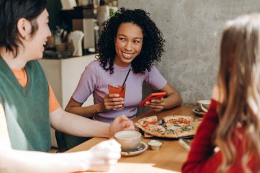 Üç neşeli genç arkadaş bir restoran masasında oturmuş pizza, içki ve hoş sohbetin tadını çıkarıyor, sıcak ve sosyal bir ortam yaratıyorlar. Yiyecek konsepti