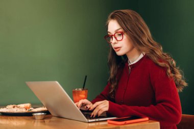 Kırmızı gözlüklü genç bir kadın ve uyumlu bir hırka rahat bir kafede dizüstü bilgisayarında çalışıyor, pizza ve meyve suyunun tadını çıkarırken görevlerine konsantre oluyor ve ambiyansın tadını çıkarıyor.