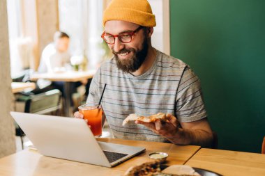 Modern, modaya uygun bir kafede çalışırken pizza yiyip meyve suyunu yudumlayan sakallı ve gözlüklü mutlu bir erkek serbest yazar, rahat ve üretken bir hayat tarzını benimsiyor.