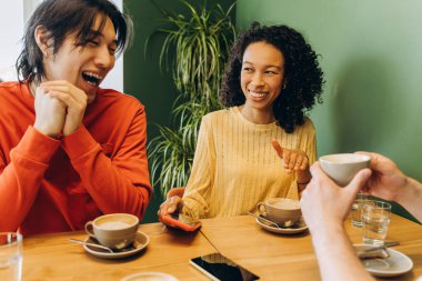 Kafe masasında oturan mutlu arkadaşlar kahvenin tadını çıkarırken gülüp sohbet ediyorlar. Keyif ve bağlantı dolu samimi ve davetkar bir ortam yaratıyorlar.