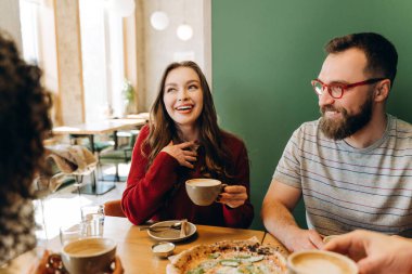 Üç neşeli arkadaş kahve içerken lezzetli bir pizzayı paylaşıyorlar, gülüyorlar ve sıcak ve güzel titreşimlerle dolu sıcak bir restoranda eğleniyorlar. İletişim konsepti