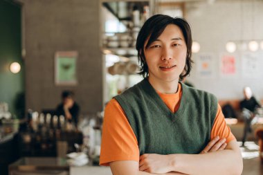 Genç Asyalı erkek barista kollarını kavuşturup gülümsüyor, turuncu tişört ve yeşil yelek giyiyor, arka planda bulanık müşterilerle modern kahve dükkanında duruyor.