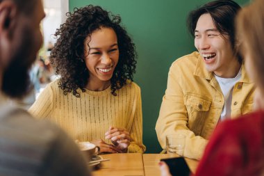 Bir grup neşeli çoklu-etnik arkadaş, modern bir kahve dükkanında masada oturup sohbet ederek ve gülerek rahatça vakit geçiriyorlar. Rahatlatıcı kavram