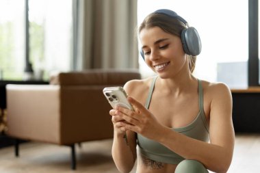 Gülümseyen sporcu kadın kulaklıkla müzik dinlerken akıllı telefonuna göz atıyor, sıcak oturma odasında yoğun bir eğitim seansından sonra hak ettiği molayı alıyor. Spor konsepti