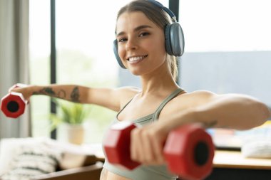 Kablosuz kulaklık takan mutlu sporcu kadın evdeki günlük antrenmanı sırasında kırmızı dambılları kaldırıyor ve antrenman seansının tadını çıkarıyor. Eğitim kavramı