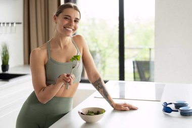 Modern bir mutfakta taze salatadan zevk alan gülümseyen sporcu kadın, sağlıklı bir yaşam tarzını ve dengeli bir diyeti kucaklarken mutluluk ve refahı yayıyor. Yiyecek konsepti