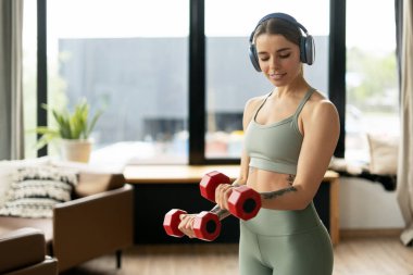 Konsantre fitness meraklısı kırmızı dambılları kaldırıp evdeki oturma odasında egzersiz yaparken kablosuz kulaklıkla müzik dinliyor.
