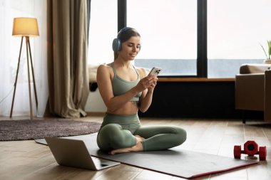 Genç sporcu kadın kulaklık takıyor ve yoga minderinde otururken akıllı telefonuyla meşgul oluyor. Etrafı dizüstü bilgisayar ve halterlerle çevrili. Çevrimiçi alışveriş kavramı