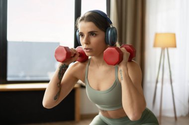 Yoğun bir şekilde konsantre olan, kulaklık takan bir sporcu kadın evde çömelme hareketleri yaparken kırmızı dambılları kaldırıyor. Etkin yaşam tarzı kavramı