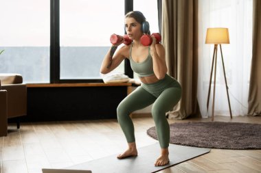 Kulaklık takan genç atletik kadın evde dizüstü bilgisayarında müzik dinlerken ve spor yaparken dambıllarla çömelme hareketleri yapıyor. Eğitim kavramı