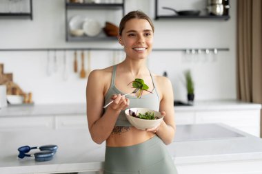 Spor kıyafetleri giyen genç bir kadın modern mutfağında sağlıklı bir salata yerken idmandan sonra dengeli bir yaşam tarzını benimsiyor. Sağlık hizmeti kavramı