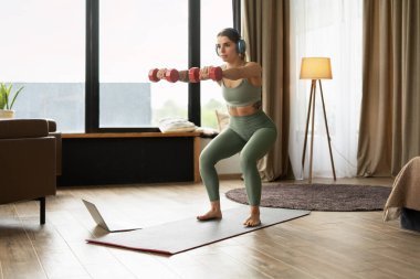 Kulaklık takan genç bir kadın, oturma odasında dizüstü bilgisayarındaki antreman dersini takip ederken dambıllarla çömeliyor. Çevrimiçi eğitim kavramı