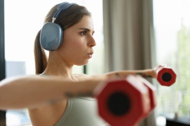 Kulaklık takan sportmen kadın sporcu evde egzersiz yaparken kırmızı dambılları kaldırıyor, dayanıklılığı ve sağlıklı yaşam tarzı ve formda kalma alışkanlığını somutlaştırıyor. Eğitim kavramı