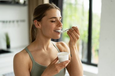 Evde antrenman yaptıktan sonra yoğurt yiyen gülümseyen sporcu kadın, sağlıklı yaşam tarzını ve kas iyileşmesi ve genel sağlık için egzersiz sonrası beslenmeyi teşvik ediyor.