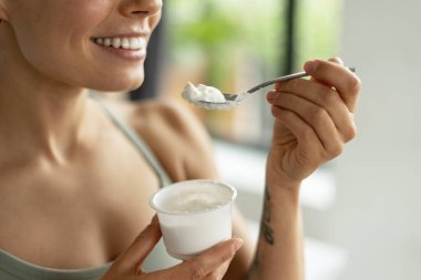 Gülümseyen kadın bir kaşık dolusu kremalı yoğurdun tadını çıkarıyor, sağlıklı beslenme alışkanlıklarını kucaklıyor ve beslenme ve sağlıkla dolu dengeli bir yaşam tarzını teşvik ediyor. Yiyecek konsepti