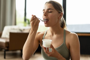 Formda bir kadın, egzersizini tamamladıktan sonra sağlıklı bir yoğurt atıştırmasından zevk alıyor, hem egzersiz hem de besleyici yeme alışkanlıklarını en iyi şekilde vurgulayan dengeli bir yaşam tarzını somutlaştırıyor. Yiyecek konsepti