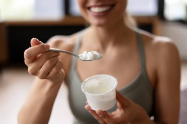 Gülümseyen bir sporcunun elinde taze yoğurt kaşığı ve idmandan sonra elinde yoğurt kabıyla sağlıklı protein zengini bir atıştırmalığın tadını çıkarırken yakın plan fotoğrafı. Yiyecek konsepti