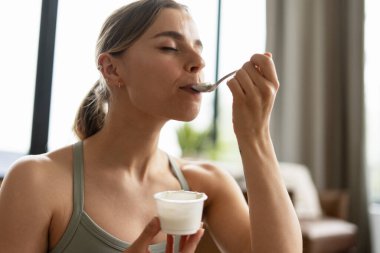Sağlıklı genç bir kadın, canlandırıcı bir egzersizin ardından besleyici bir yoğurdun tadını çıkarıyor, dengeli bir yaşam tarzını somutlaştırıyor ve iyileşme için egzersiz sonrası beslenmenin önemini vurguluyor.