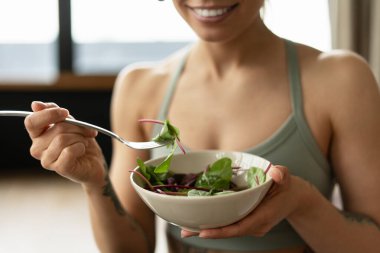 Açık yeşil spor sutyen giyen, elinde çatalla bir kase taze salata tutan, besleyici egzersiz sonrası yemeğin tadını çıkaran, gülümseyen sporcu kadın. Sağlıklı yaşam tarzı kavramı