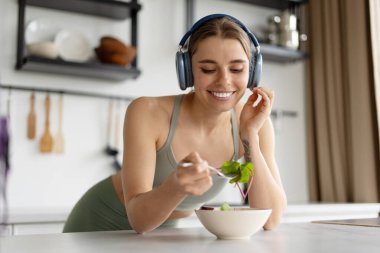 Gülümseyen sporcu kadın kulaklık takıyor ve mutfakta sağlıklı yiyeceklerin tadını çıkarıyor, müzik dinliyor ve antrenmandan sonra taze sebze salatası yiyor, sağlıklı yaşam tarzını ve dengeli beslenme düzenini destekliyor.
