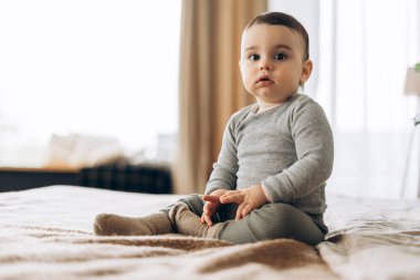 Sevimli bebek rahat bir yatak odasında rahat bir yatakta oturuyor, yumuşak giysiler giyiyor ve merakla etrafı izliyor, huzur dolu bir sabah ışığında masumiyet ve neşe saçıyor.