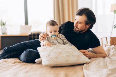 Baba ve bebek birlikte kaliteli zaman geçiriyorlar, yatakta uzanıp yumuşak bir yastıkla etkileşime geçiyorlar, aile bağlarının iç açıcı bir sahnesini yaratıyorlar. Babalık kavramı