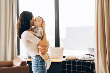 Genç anne bebeğini kollarında öpüyor oturma odasındaki pencerenin yanında duruyor şefkatli bir anın ve anneliğin tadını çıkarıyor.