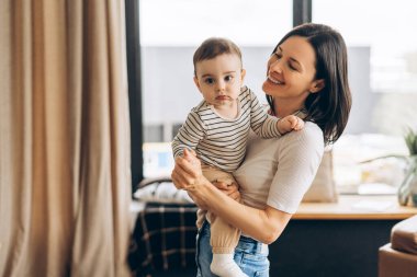 Gülümseyen bir anne, bebeğini kollarında tutarak eve davet edilmelerinin sıcaklığının ve rahatlığının tadını çıkarırken, sevgi ve sevgi dolu bir anı kutluyor. Annelik kavramı